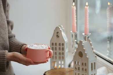 Photo of Woman holding cup of delicious drink near windowsill with house shaped lanterns indoors, closeup. Space for text