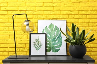 Photo of Beautiful plant in pot, lamp and pictures on black table near yellow brick wall. Interior accessories