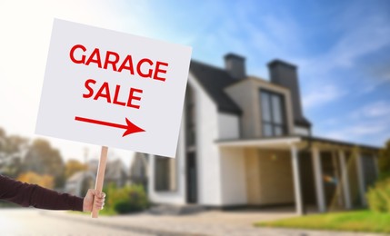 Image of Man holding sign with text GARAGE SALE and blurred view of modern house