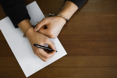 Criminal in handcuffs writing confession at desk, top view