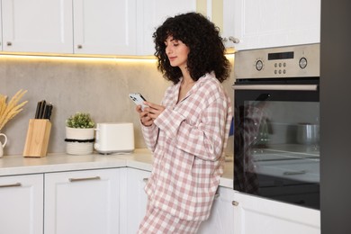 Beautiful young woman in stylish pyjama using smartphone in kitchen