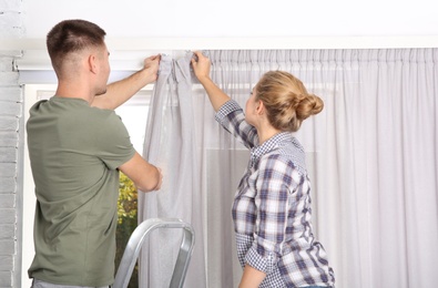 Young couple hanging window curtain indoors. Interior decor element