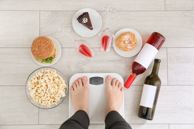 Photo of Woman standing on scales surrounded by different food and alcohol after party indoors, top view