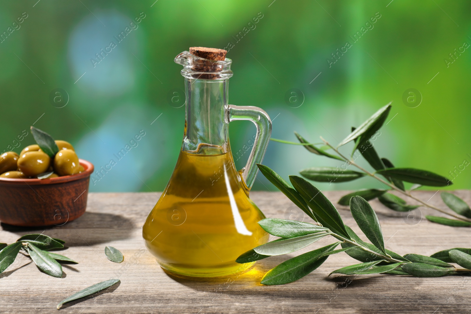 Photo of Jug of cooking oil, olives and green leaves on wooden table against blurred background