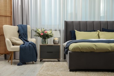 Large comfortable bed, armchair and beautiful bouquet on bedside table in room. Bedroom interior