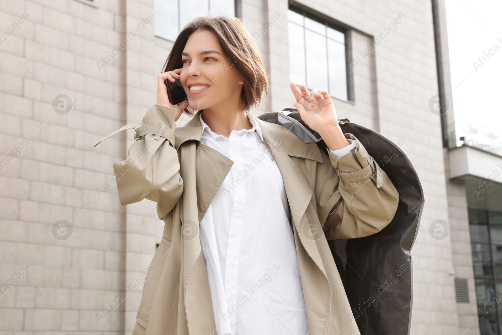 Photo of Woman holding garment cover with clothes while talking on phone outdoors. Dry-cleaning service