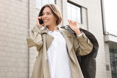 Woman holding garment cover with clothes while talking on phone outdoors. Dry-cleaning service
