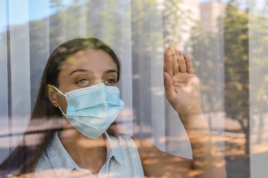 Photo of Stressed woman in protective mask looking out of window, view through glass. Self-isolation during COVID-19 pandemic