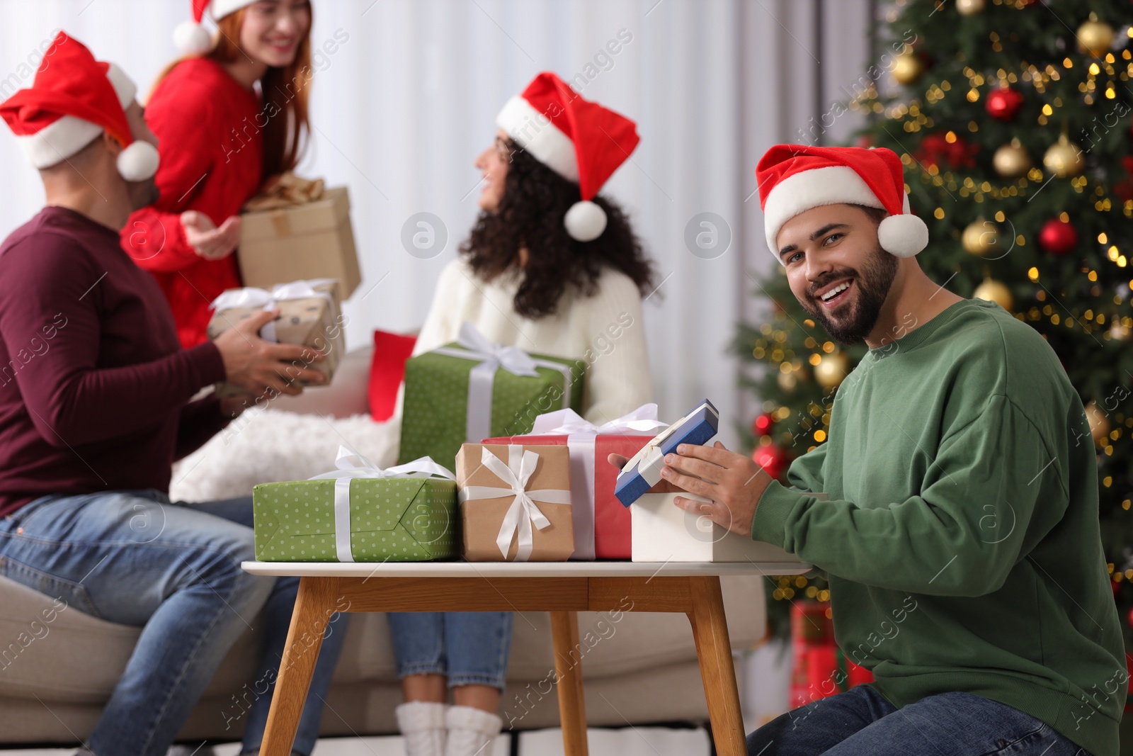 Photo of Christmas celebration in circle of friends. Happy young man opening gift at home, selective focus