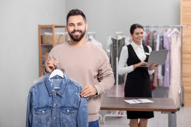 Photo of Dry-cleaning service. Happy man holding hanger with denim jacket in plastic bag indoors. Worker using laptop at workplace
