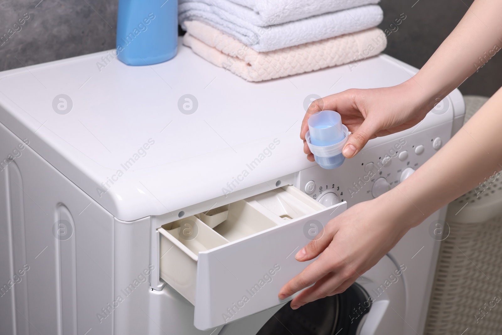 Photo of Woman pouring fabric softener from cap into washing machine, closeup