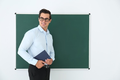 Photo of Young teacher with book near blank chalkboard in classroom. Space for text