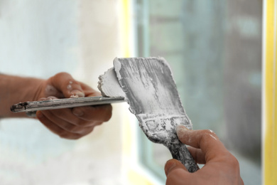 Professional worker with putty knifes and plaster near window indoors, closeup. Interior repair