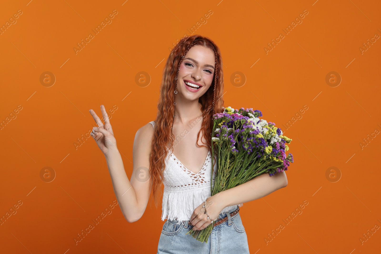 Photo of Beautiful young hippie woman with bouquet of colorful flowers showing V-sign on orange background