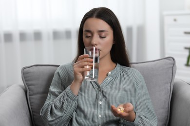 Depressed woman with glass of water taking antidepressant pills in armchair indoors