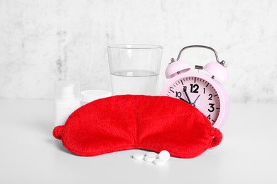 Photo of Red sleep mask, glass of water, pills and alarm clock on white table