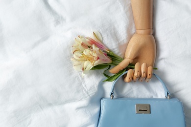 Photo of Stylish bag with spring flowers and wooden mannequin hand on crumpled sheet, flat lay. Space for text