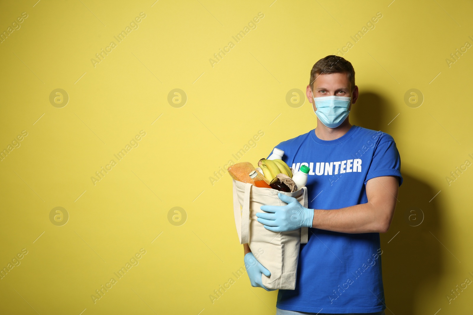 Photo of Male volunteer in protective mask and gloves with products on yellow background, space for text. Aid during coronavirus quarantine