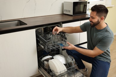 Photo of Smiling man loading dishwasher with glasses in kitchen