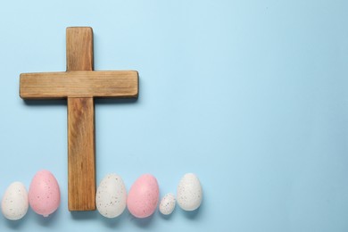 Wooden cross and painted Easter eggs on light blue background, flat lay. Space for text