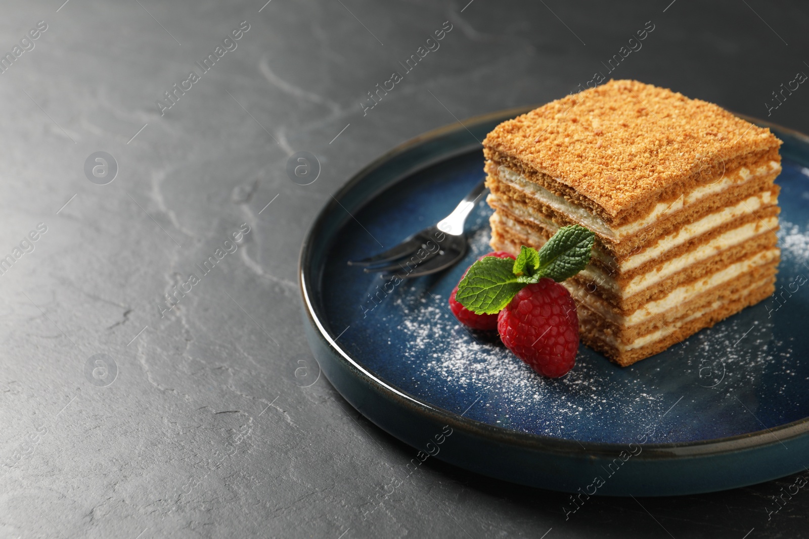 Photo of Slice of delicious layered honey cake with mint, raspberries and powdered sugar served on black table. Space for text