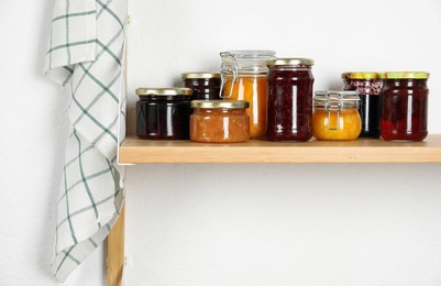 Photo of Jars with different homemade preserves on wooden shelf near white wall