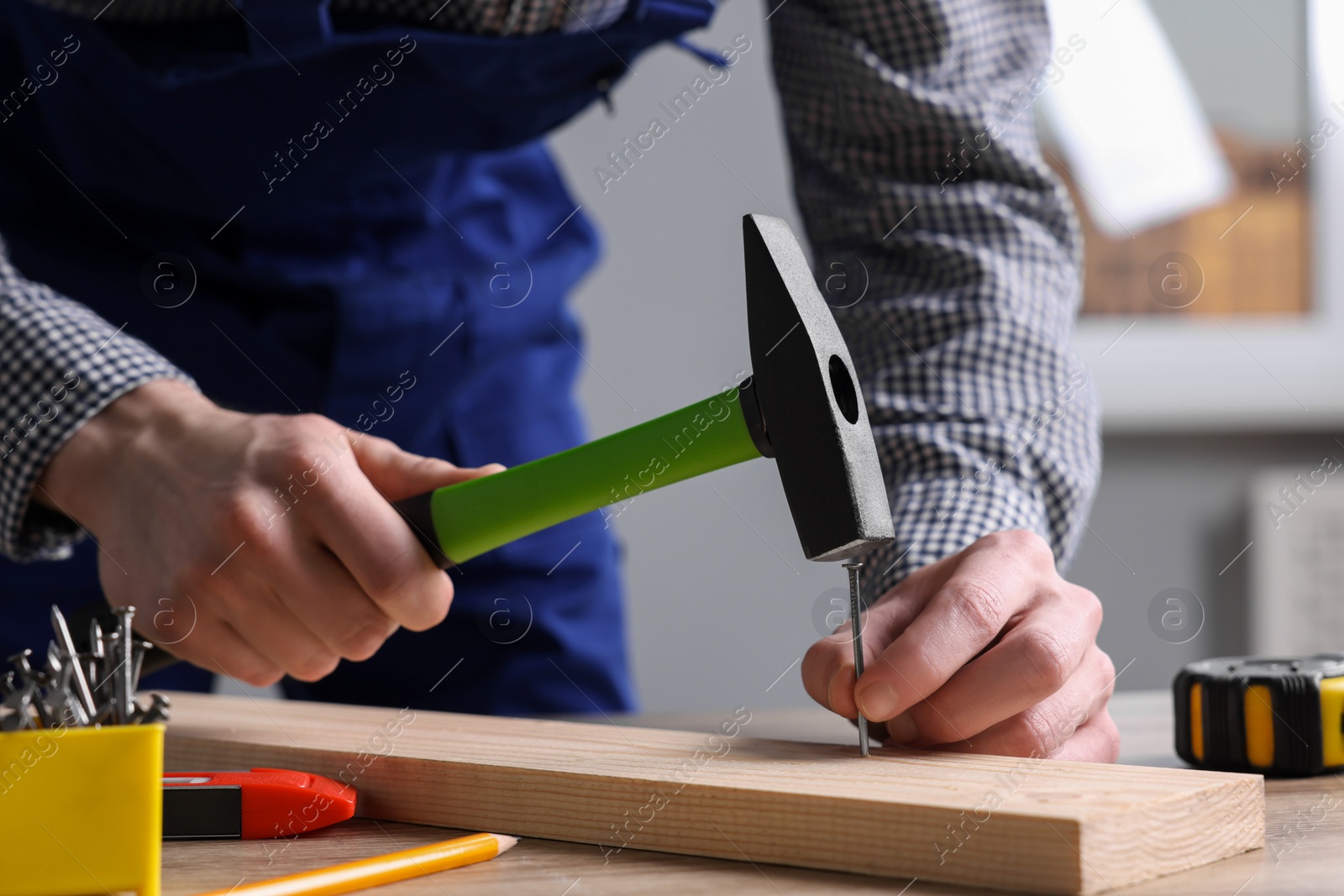 Photo of Professional repairman hammering nail into board at wooden table indoors, closeup