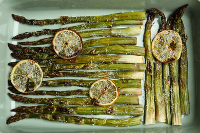 Photo of Oven baked asparagus with lemon slices in ceramic dish, flat lay