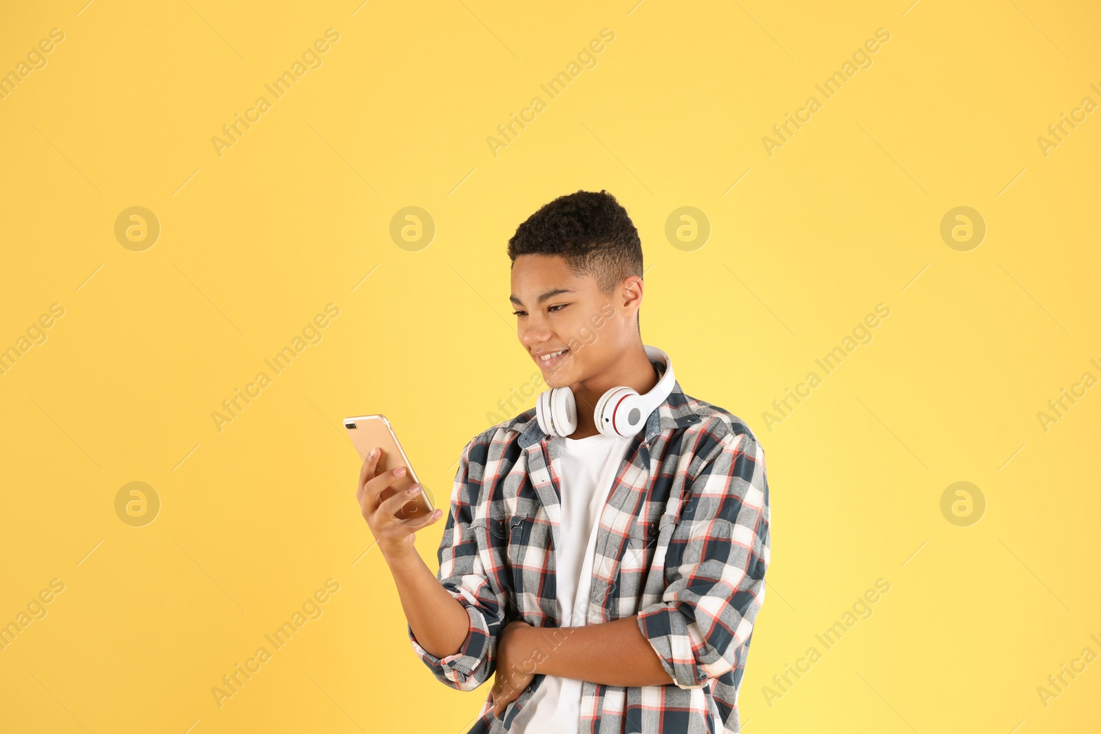 Photo of African-American teenage boy with headphones using mobile phone on color background