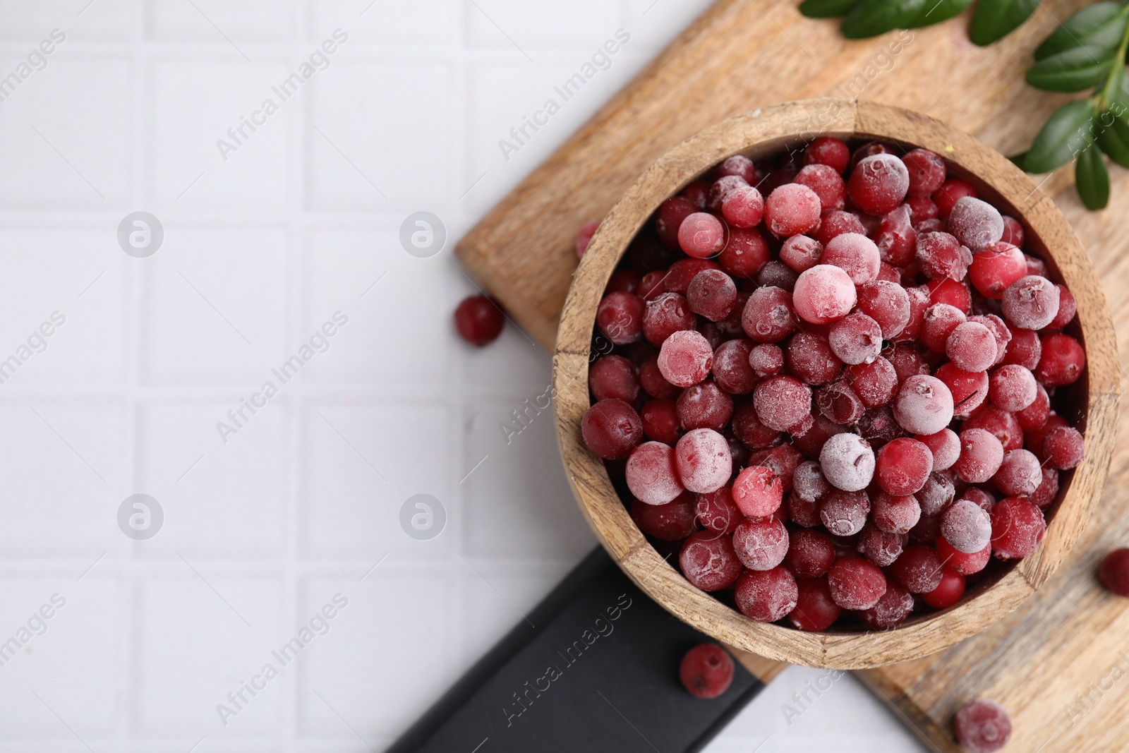 Photo of Frozen red cranberries in bowl and green leaves on white tiled table, top view. Space for text