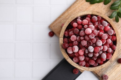 Photo of Frozen red cranberries in bowl and green leaves on white tiled table, top view. Space for text