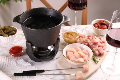 Photo of Fondue pot with oil, forks, raw meat pieces, glasses of red wine and other products on white marble table