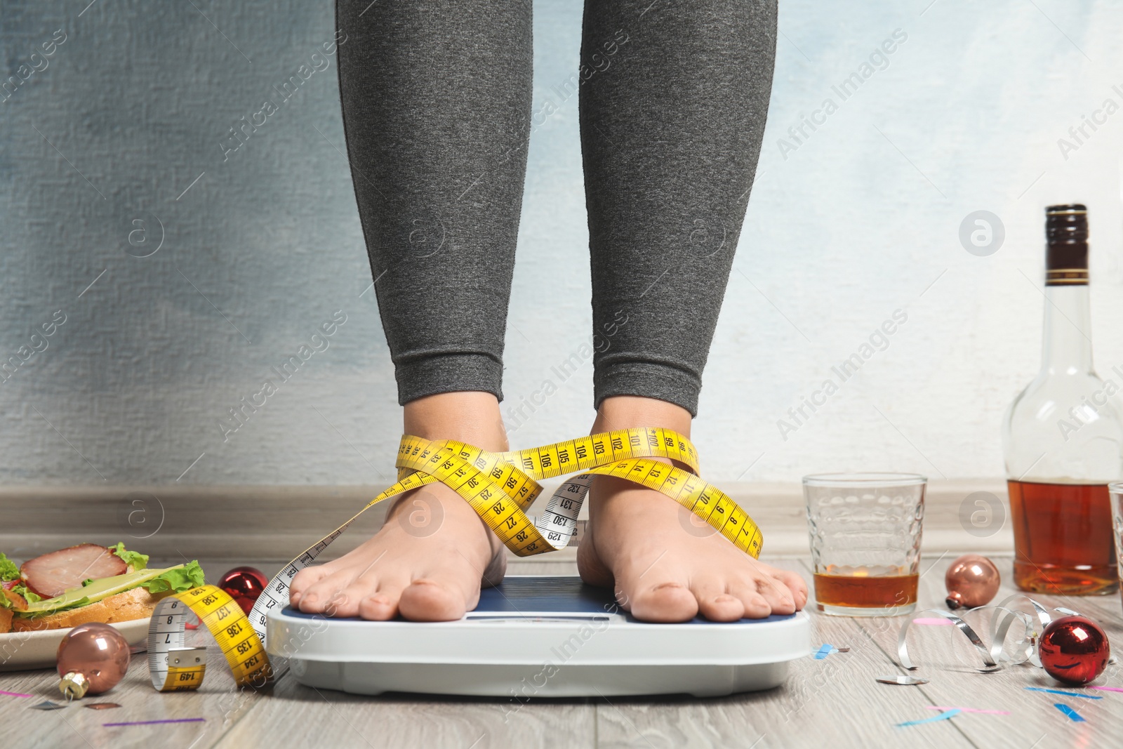 Photo of Woman with measuring tape using scale surrounded by food and alcohol after party on floor. Overweight problem