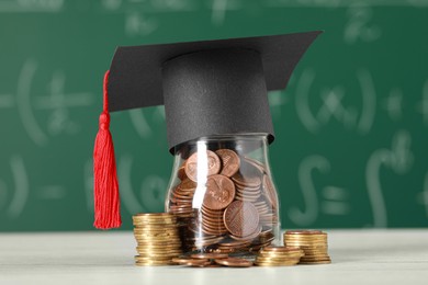 Photo of Scholarship concept. Glass jar with coins and graduation cap on white wooden table