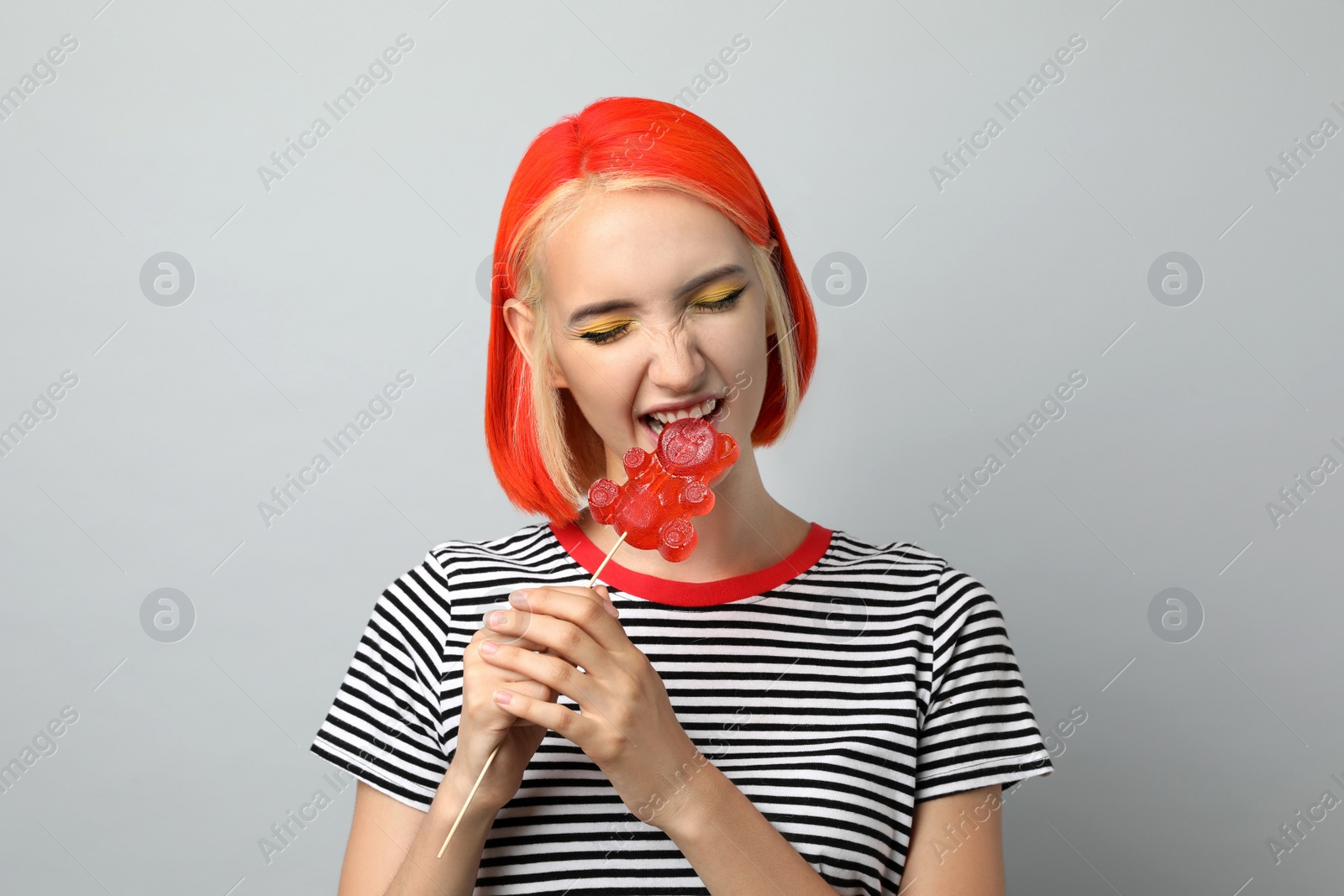 Photo of Beautiful young woman with bright dyed hair eating bear shaped candy on light grey background