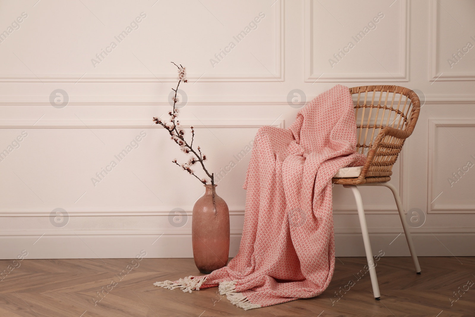 Photo of Flowering tree twig in glass vase on floor near chair with blanket at white wall