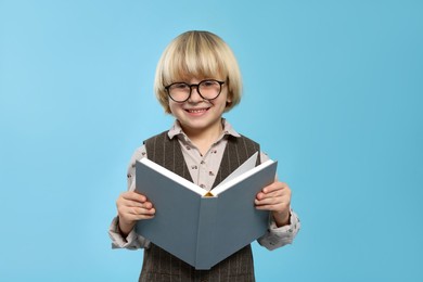 Cute little boy in glasses reading book on light blue background