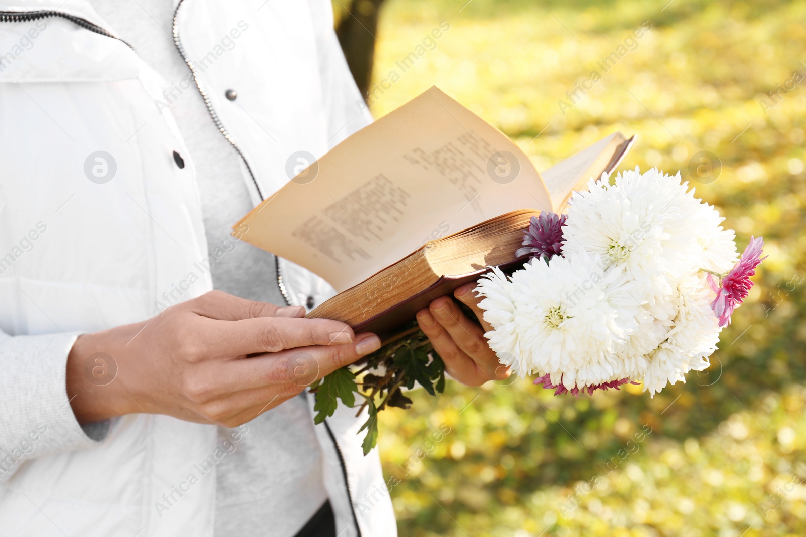 Photo of Woman reading book and holding beautiful flowers outdoors on autumn day, closeup