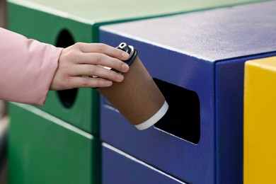 Photo of Woman throwing paper coffee cup into garbage bin outdoors, closeup. Waste sorting