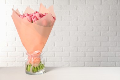 Bouquet of beautiful pink tulips in vase on white table near brick wall, space for text