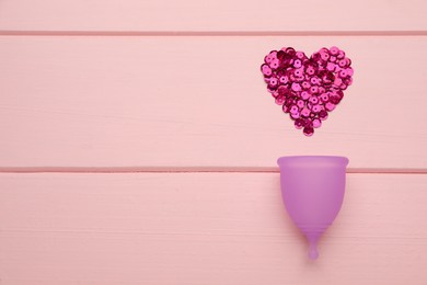 Menstrual cup near heart made of sequins on pink wooden background, flat lay. Space for text