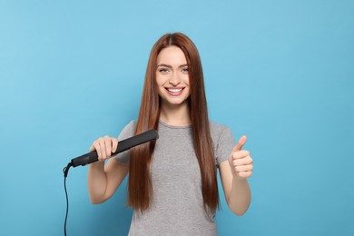Beautiful woman using hair iron and showing thumbs up on light blue background