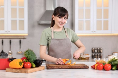 Photo of Happy young housewife cutting bell pepper at white marble table in kitchen