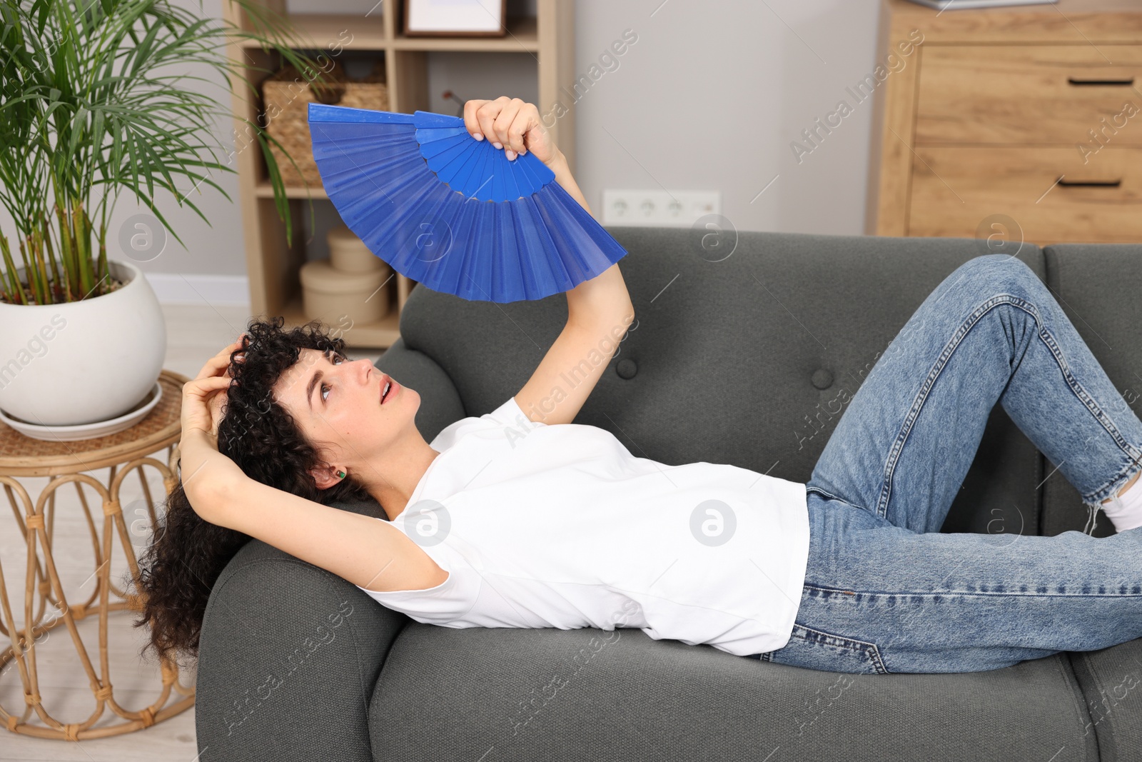 Photo of Young woman waving blue hand fan to cool herself on sofa at home