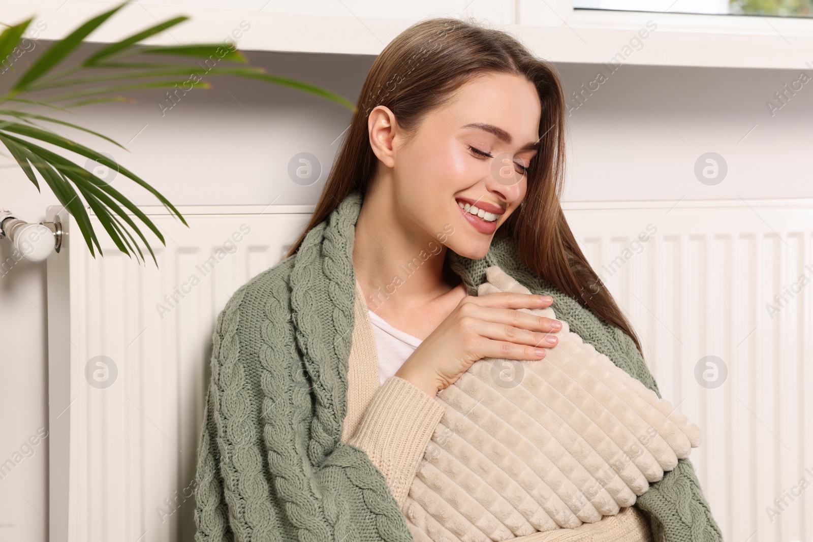 Photo of Woman with blanket hugging pillow near heating radiator indoors