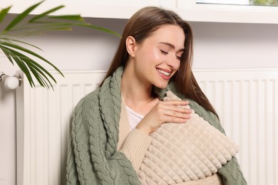 Photo of Woman with blanket hugging pillow near heating radiator indoors