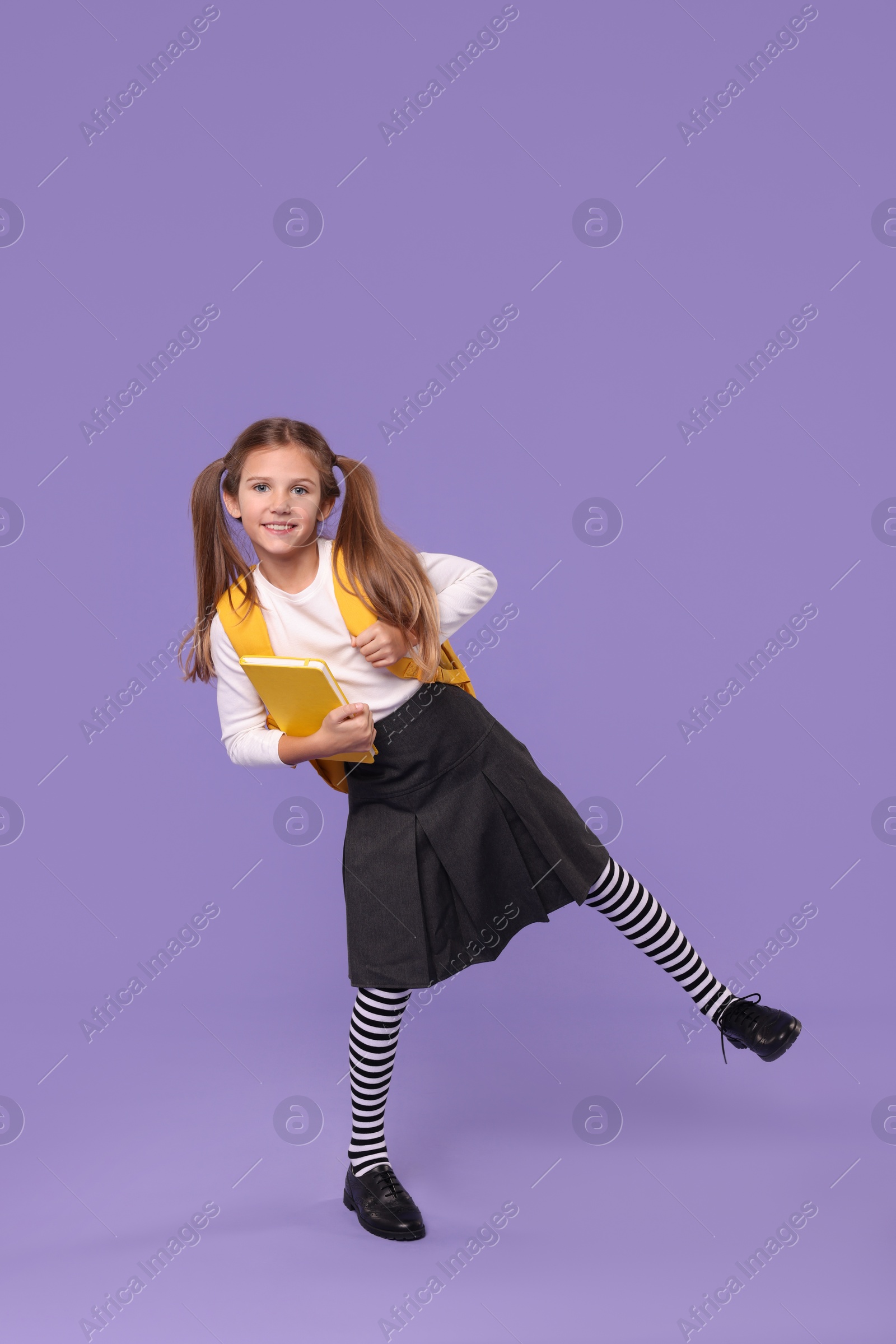 Photo of Smiling schoolgirl with backpack and book on violet background