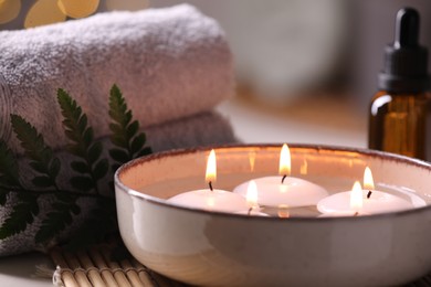 Photo of Spa composition. Burning candles in bowl, towels and bottle of essential oil on table, closeup