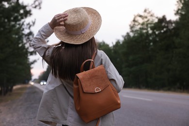 Young woman with stylish backpack near empty road in forest, back view. Space for text