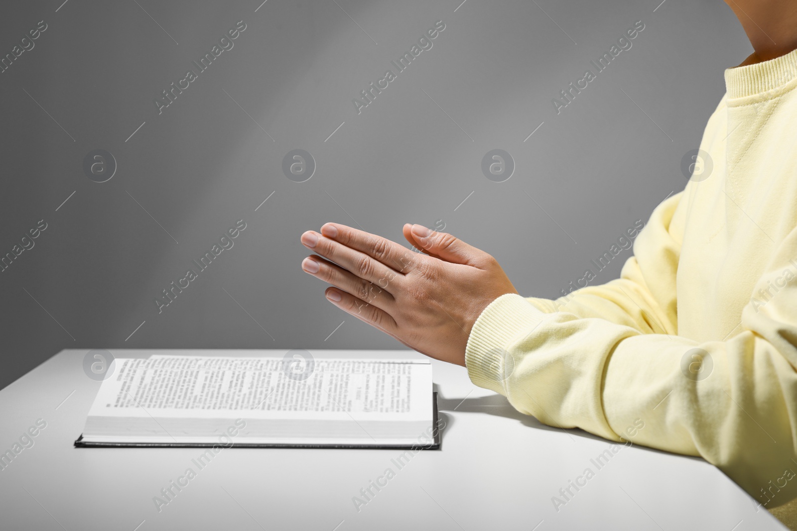 Photo of Woman praying over Bible at white table against grey background, closeup